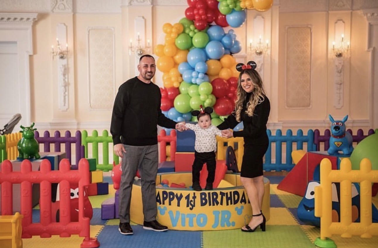 Family celebrating Vito Jr’s first birthday in a colorful indoor play area with balloons, toys, and a decorated birthday platform.”